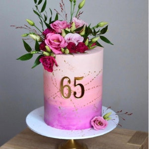 May include: A pink and purple ombre cake decorated with gold accents and a floral arrangement. The cake has the number "65" on the front.