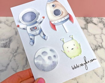 May include: A set of four space-themed stickers featuring an astronaut, a rocket ship, a moon, and an alien. The stickers are printed on white paper with a watercolour effect.