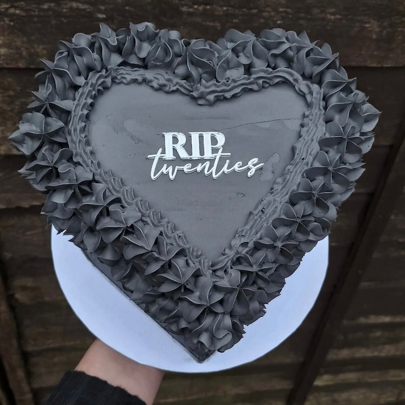 May include: A black heart-shaped cake with gray frosting and the words "RIP twenties" written in white icing.