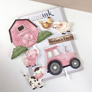May include: A set of pink and white farm-themed cake toppers. The set includes a red barn, a pig, a cow, a sheep, a tractor, a chicken, and a sign that says "Stefan's Farm".