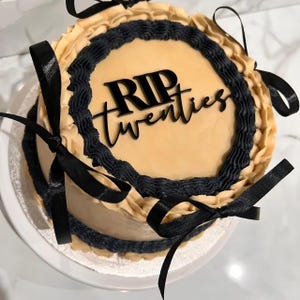May include: A beige cake with black frosting and a black ribbon. The cake is decorated with the words "RIP Twenties" in black lettering.