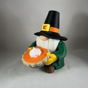 Könnte beinhalten: Eine Thanksgiving-Wichtelfigur, die einen Kürbiskuchen mit Schlagsahne hält. Der Wichtel trägt einen schwarzen Hut mit orangefarbenem Band und gelber Schnalle, einen grünen Mantel, gelbe Handschuhe und einen weißen Bart. Der Kuchen hat eine orange Füllung und einen braunen Boden.