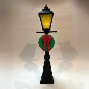Op de afbeelding: Een zwarte decoratieve lantaarnpaal met een groene krans en rode strik. De lamp heeft een geel licht en een puntige top. De lantaarnpaal is een feestelijke vakantieversiering.