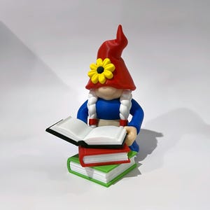 May include: A figurine of a gnome wearing a red hat with a yellow flower and white braided hair, dressed in a blue shirt. The gnome is holding an open book while sitting on a stack of two closed books, one red and one green, against a white background.