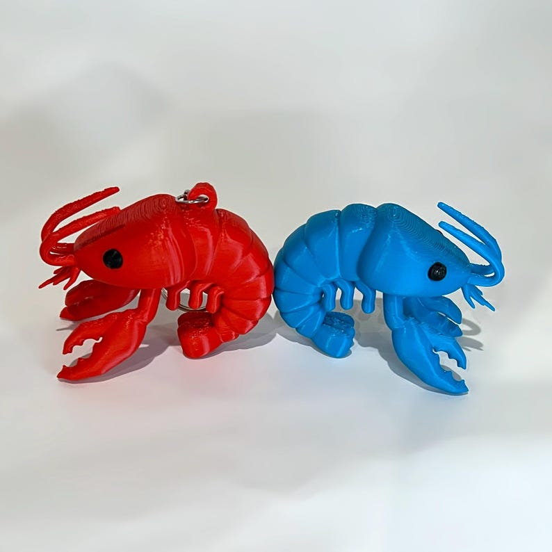 May include: Two 3D-printed lobster figurines. One is bright red, and the other is a vibrant blue. Each lobster has black eyes and a keychain loop. The claws and antennae are detailed. The figurines are on a white background.