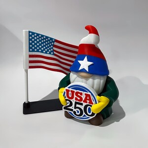May include: A patriotic gnome figurine wearing a red, white, and blue hat and green jacket, holding a sign that says "USA 250." An American flag waves in the background. The gnome has a white beard and yellow gloves.
