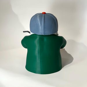 May include: A green figurine of a person wearing a blue baseball cap with a red button on top and a green jacket. The figurine is viewed from the back, with the person holding a black object.