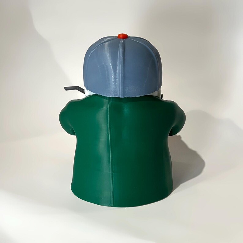 May include: A green figurine of a person wearing a blue baseball cap with a red button on top and a green jacket. The figurine is viewed from the back, with the person holding a black object.