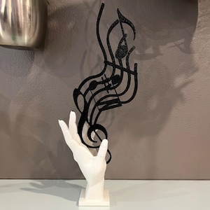 Könnte beinhalten: Eine weiße Handskulptur, die ein schwarzes Musiknoten-Design aus Metall hält. Die Skulptur steht auf einem weißen Sockel.