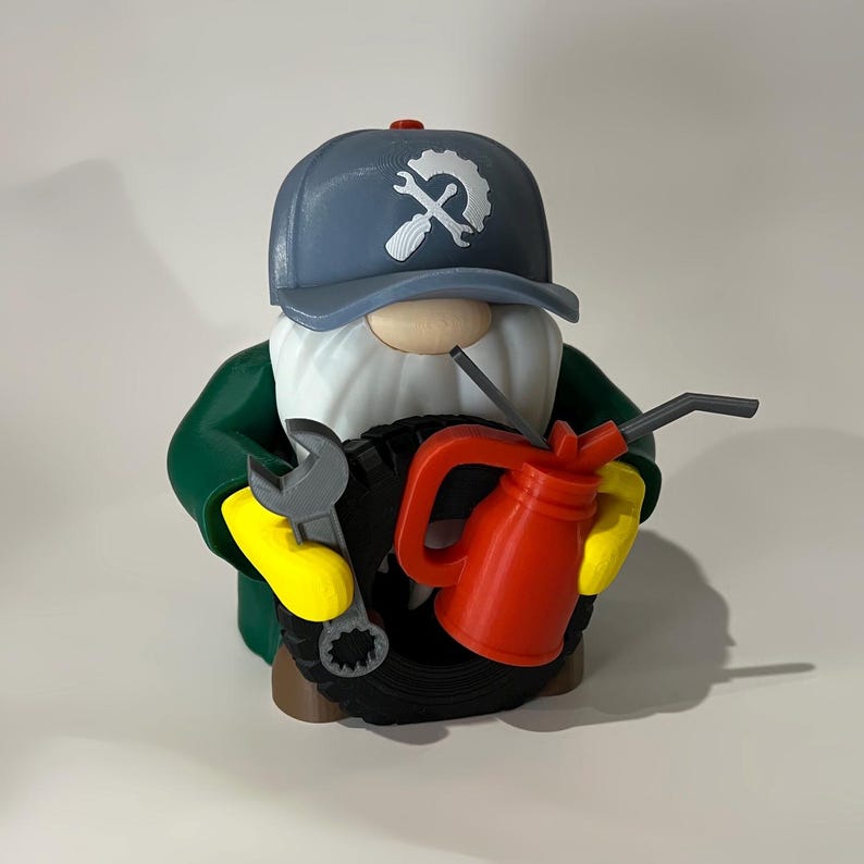 May include: A whimsical gnome figurine dressed as a mechanic, wearing a blue cap with a wrench logo. The gnome has a white beard, green jacket, yellow gloves, and holds a wrench and a red oil can. A black tire is in the center.