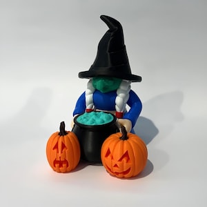 May include: A Halloween-themed figurine of a witch with a black hat, green face, and blue dress, holding a black cauldron with turquoise liquid. Two orange jack-o'-lanterns flank the witch, creating a festive display.