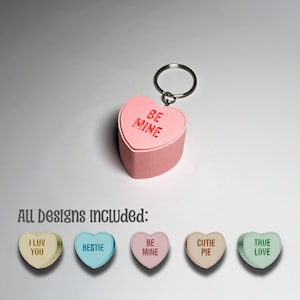 Puede incluir: Un llavero rosa en forma de corazón con las palabras "BE MINE" grabadas en la parte superior. El llavero incluye un anillo plateado. Debajo, la imagen muestra otros diseños en forma de corazón con frases como "I LUV YOU" y "BESTIE".