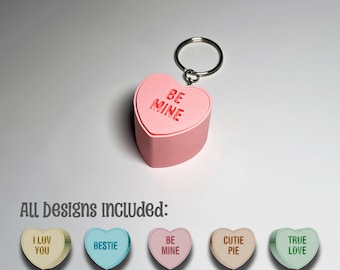 May include: A pink heart-shaped keyring with the words "BE MINE" engraved on the top. The keyring includes a silver ring. Below, the image shows other heart-shaped designs with phrases like "I LUV YOU" and "BESTIE".