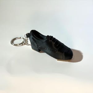 Puede incluir: Un llavero de zapato de claqué negro en miniatura. El zapato pequeño tiene tacón y puntera, y está hecho para parecerse a un zapato de claqué real. El llavero es un artículo de novedad.