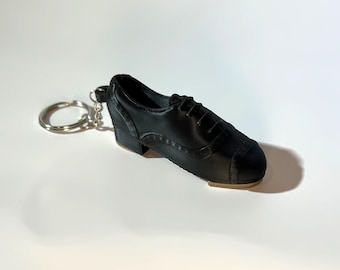 May include: A black miniature tap shoe keychain with a silver key ring. The small shoe has a heel and a toe tap, and is made to look like a real tap shoe. The keychain is a novelty item.