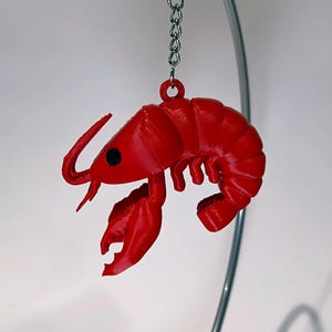 May include: A vibrant red lobster keyring, hanging from a silver chain. The lobster features detailed claws, antennae, and a segmented body. The keyring is a novelty item, possibly 3D-printed, set against a white backdrop.