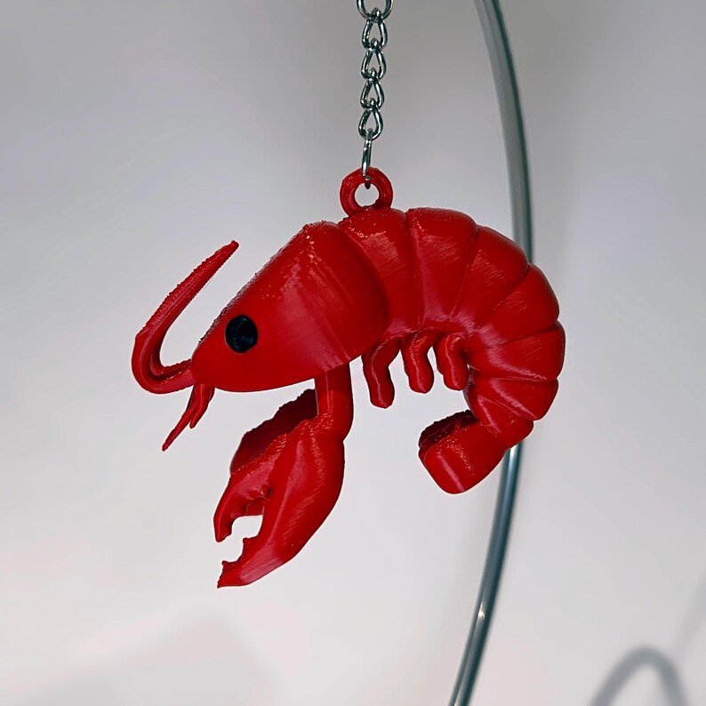 May include: A vibrant red lobster keyring, hanging from a silver chain. The lobster features detailed claws, antennae, and a segmented body. The keyring is a novelty item, possibly 3D-printed, set against a white backdrop.