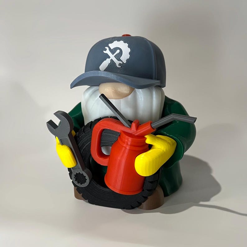May include: A whimsical gnome figurine dressed as a mechanic, wearing a blue cap with a wrench logo. The gnome holds a wrench, a red oil can, and a tire. The gnome's outfit includes a green jacket and yellow gloves.