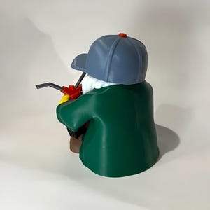 May include: A 3D-printed figurine of a person wearing a blue baseball cap with a red button on top, a green jacket, and a white beard. The figurine is holding a red and black tool.