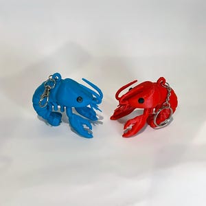 May include: Two lobster keyrings, one blue and one red, are shown against a white backdrop. Each keyring features a lobster design with a silver-coloured clasp and ring. The lobsters have black eyes and detailed claws.