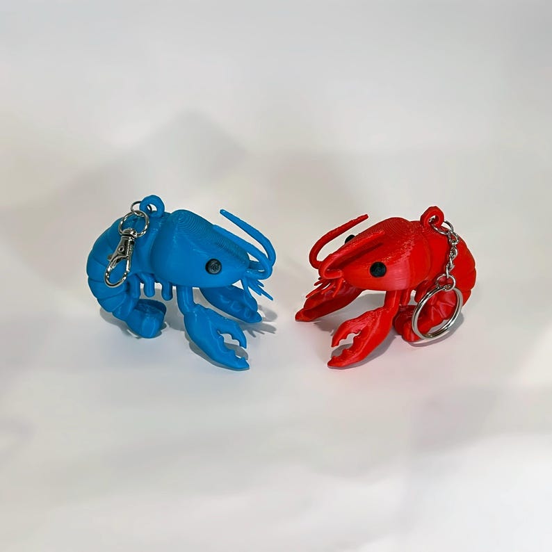 May include: Two lobster keyrings, one blue and one red, are shown against a white backdrop. Each keyring features a lobster design with a silver-coloured clasp and ring. The lobsters have black eyes and detailed claws.