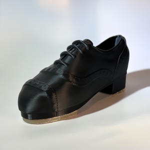 May include: Black tap shoe with a low heel and a leather-like appearance. The shoe features a rounded toe, decorative stitching, and a tap plate on the sole. The laces are black. This shoe is designed for tap dancing.