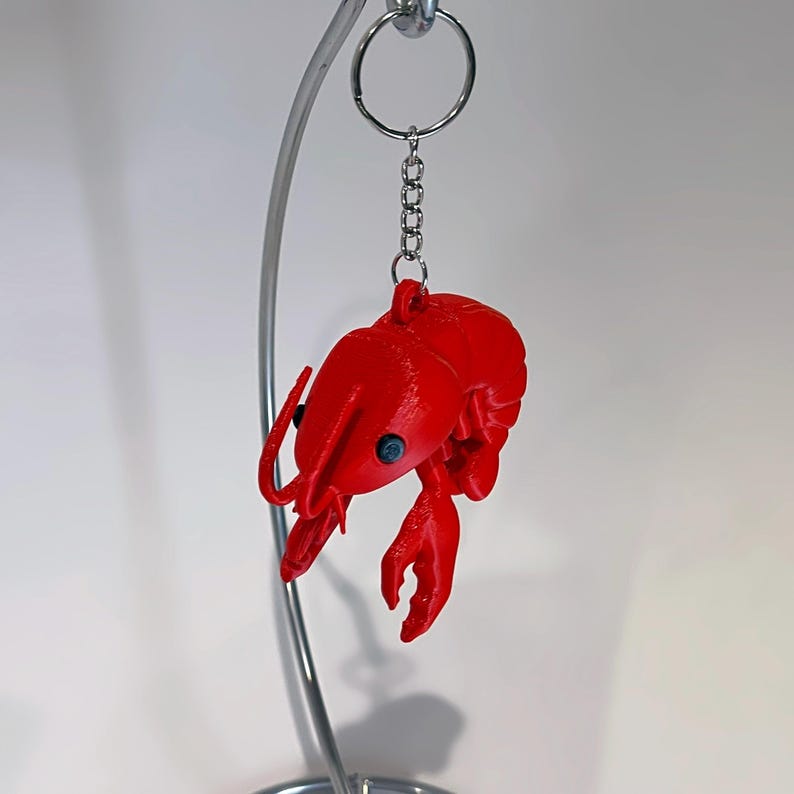 May include: A vibrant red lobster keyring with a silver chain and ring. The lobster features detailed claws, antennae, and small blue eyes. The keyring is suspended against a white backdrop.