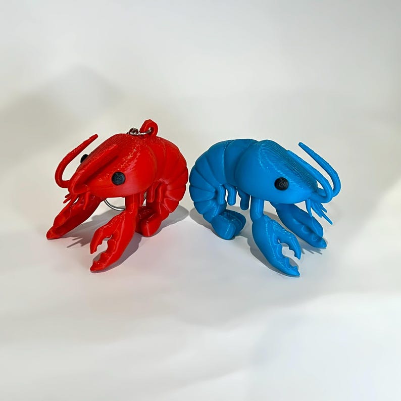 May include: Two 3D-printed lobster figurines. One is bright red, the other is a vibrant blue. Both have black eyes and detailed claw and body features. The red lobster has a small silver keychain ring attached. These decorative items are ideal for nautical-themed decor.