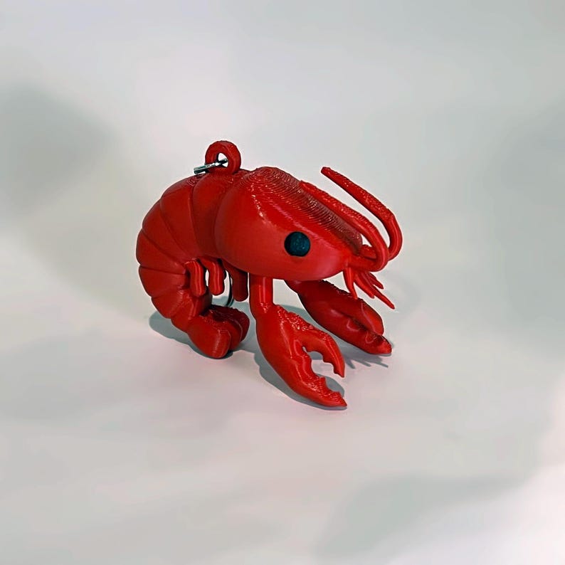 May include: A vibrant red lobster ornament with a keyring attachment. The lobster features detailed claws, antennae, and a segmented body. The ornament has a single blue eye and is likely plastic.
