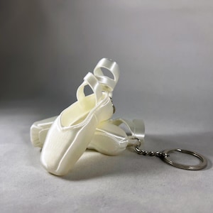 May include: A white satin ballet shoe keychain with a silver key ring. The shoe is a miniature replica of a real ballet shoe.