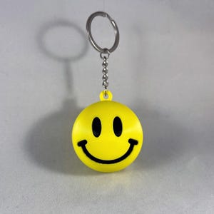 Puede incluir: Un llavero amarillo con una cara sonriente y una sonrisa y ojos negros. El llavero tiene una cadena y un anillo plateados. La cara sonriente tiene forma de esfera. El fondo es de color gris claro.