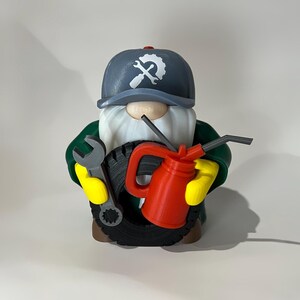 May include: A whimsical gnome figurine dressed as a mechanic, wearing a gray cap with a wrench logo. The gnome holds a tire, a wrench, and a red oil can, with yellow gloves and a white beard. The figurine is a fun decorative item.