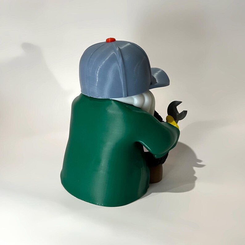 May include: A 3D-printed figurine of a person wearing a gray baseball cap with a red button, a green jacket, and holding a wrench. The figurine has a white beard and is facing away from the viewer.