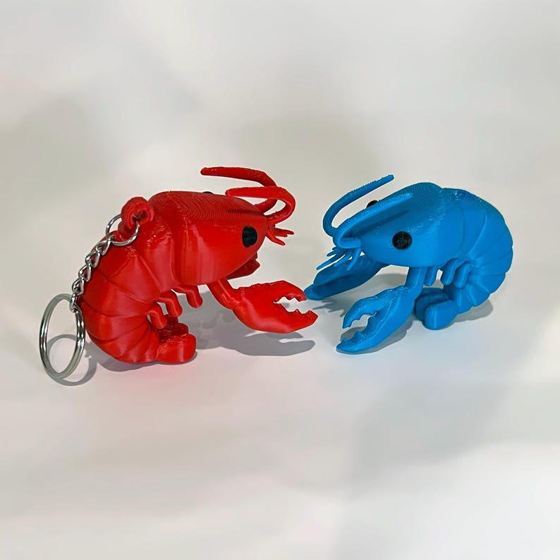 May include: A red lobster keychain and a blue lobster figurine. The red lobster has a silver chain and keyring. Both are 3D-printed with detailed claws, antennae, and body segments. The background is plain white.