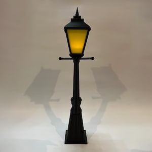 Könnte beinhalten: Eine schwarze Straßenlaterne im Vintage-Stil mit gelbem Licht. Die Lampe hat einen quadratischen Sockel, einen hohen, verzierten Pfosten und einen laternenförmigen Aufsatz. Die Höhe der Lampe beträgt ca. 152 cm.