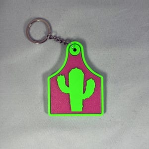 May include: A pink and green plastic keychain shaped like a cattle ear tag with a green cactus design.