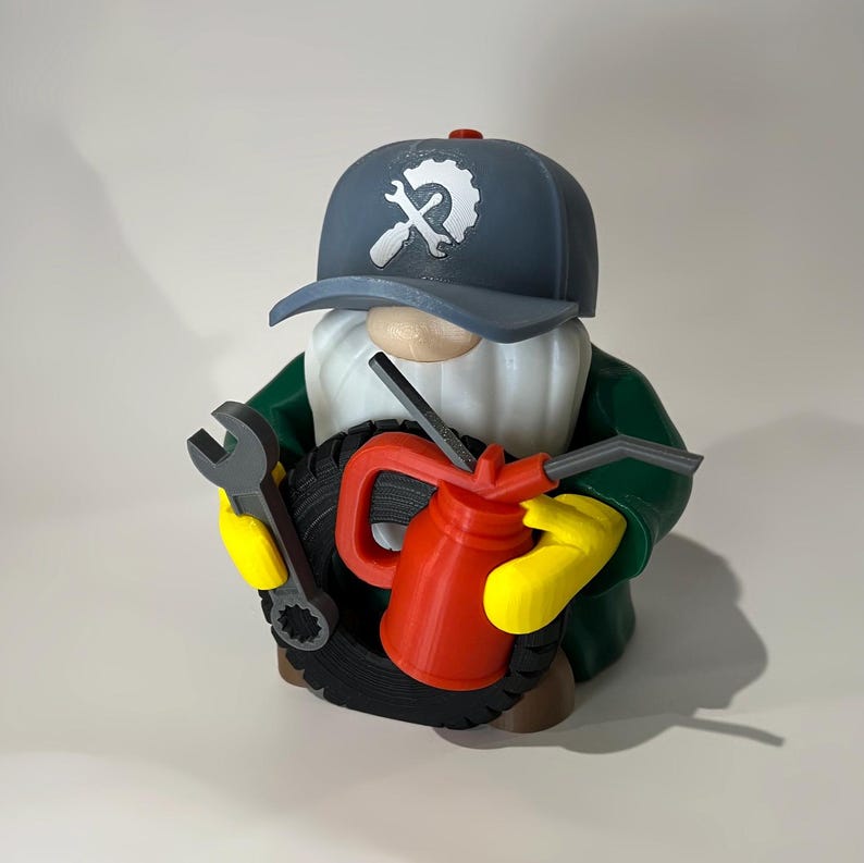 May include: A whimsical gnome figurine dressed as a mechanic, wearing a blue cap with a wrench and gear logo. The gnome holds a tire, a wrench, and a red oil can, with yellow gloves and a green jacket. The figurine is approximately 6 inches tall.