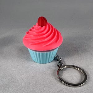 Puede incluir: Un llavero de cupcake rosa y azul con una cereza roja en la parte superior. El cupcake está hecho de plástico y tiene un diseño de espiral en la cobertura.