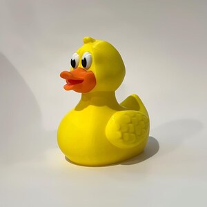 May include: A bright yellow rubber duck toy featuring a vibrant orange beak and large white eyes with black pupils. The duck has a smooth, rounded body with moulded wing details on the sides and a small tuft of feathers on the top of its head.