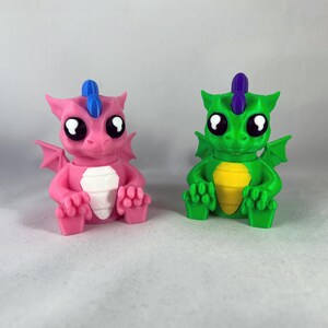 Könnte beinhalten: Zwei kleine, 3D-gedruckte Drachenfiguren. Eine ist rosa mit einem blauen Horn und die andere ist grün mit einem violetten Horn. Beide haben große Augen und sitzen mit ausgebreiteten Flügeln.