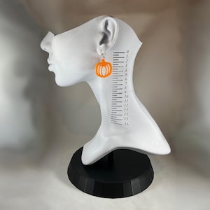 May include: Orange pumpkin earrings with silver hooks are displayed on a white mannequin head. The earrings are a festive accessory, perfect for Halloween or autumn. The mannequin has a black base and a ruler printed on the side.