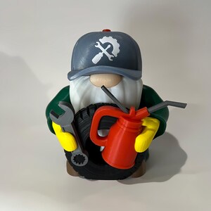 May include: A mechanic gnome figurine wearing a gray cap with a white gear logo, green shirt, and yellow gloves. The gnome holds a wrench, a tire, and a red oil can. The figurine has a long white beard and a brown base.