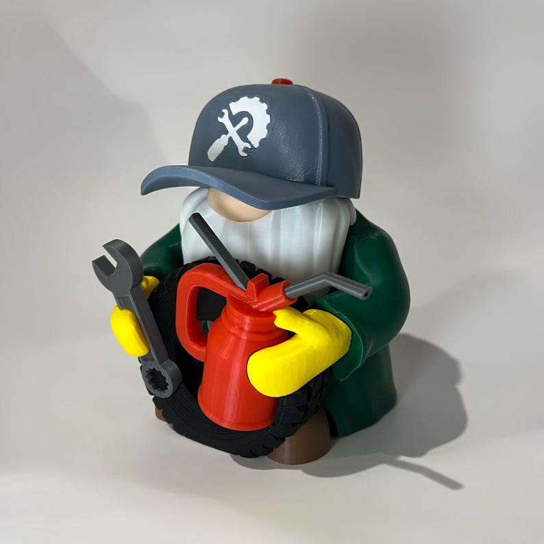 May include: A figurine of a mechanic gnome wearing a blue cap with a wrench logo, green jacket, and yellow gloves. The gnome holds a wrench and a red oil can, and is standing behind a black tire.