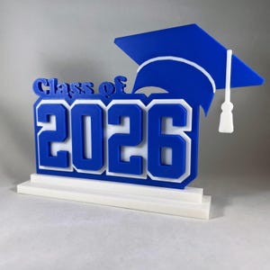 May include: A blue and white graduation decoration with the text "Class of 2026" and a graduation cap. The numbers and letters are in a bold, blocky font. The graduation cap is blue with a white tassel.