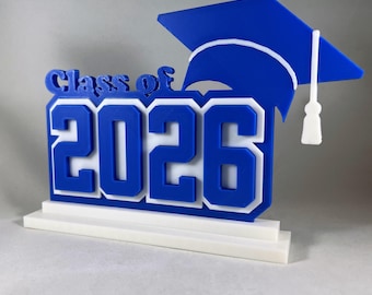 May include: A blue and white graduation decoration featuring the text "Class of 2026" and a graduation cap. The letters and numbers are in a bold font. The graduation cap is blue with a white tassel.
