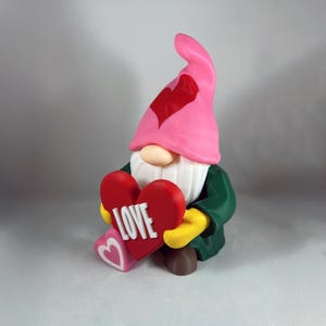 May include: A Valentine's Day gnome figurine with a pink hat featuring a red heart. The gnome wears a green jacket, yellow gloves, and holds a red heart with the word "LOVE" in white. A smaller heart with a pink heart is at its feet.