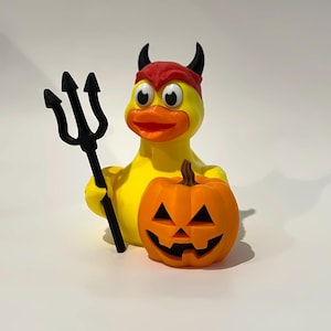 May include: A yellow rubber duck figure wearing a red headband with black devil horns. The duck holds a black trident in one wing and stands next to an orange jack-o'-lantern pumpkin with a carved face featuring black eyes, nose, and mouth.