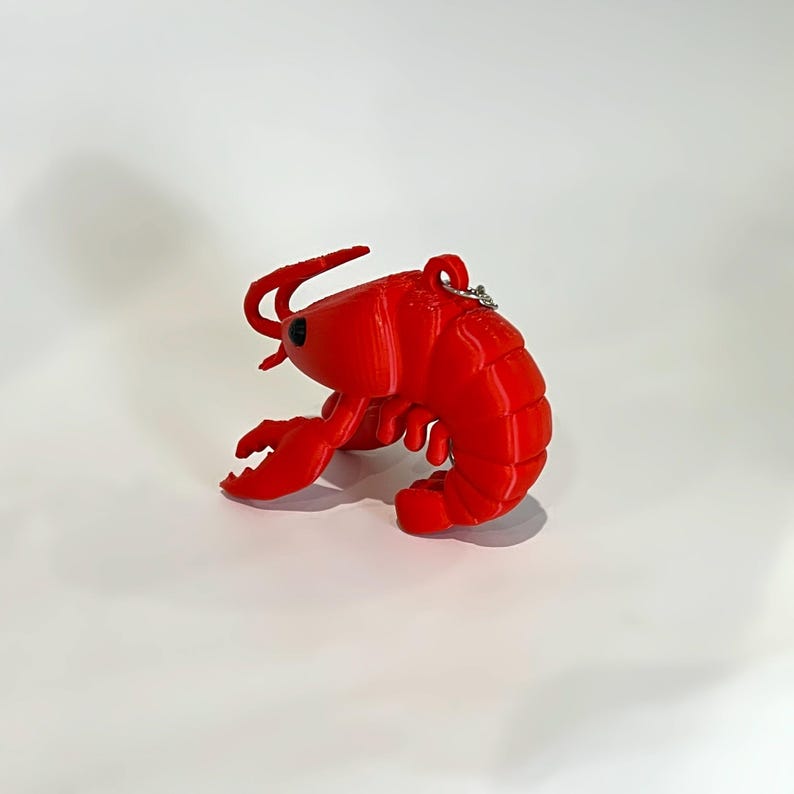 May include: A vibrant red lobster ornament with a small silver ring attached. The lobster features detailed claws, antennae, and a segmented body. The ornament is made of a solid material and is set against a white background.