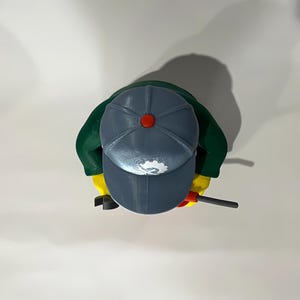 May include: Overhead view of a Lego figure wearing a gray cap with a red button, green jacket, and yellow hands. The figure holds a black and red tool. The cap has a white logo.