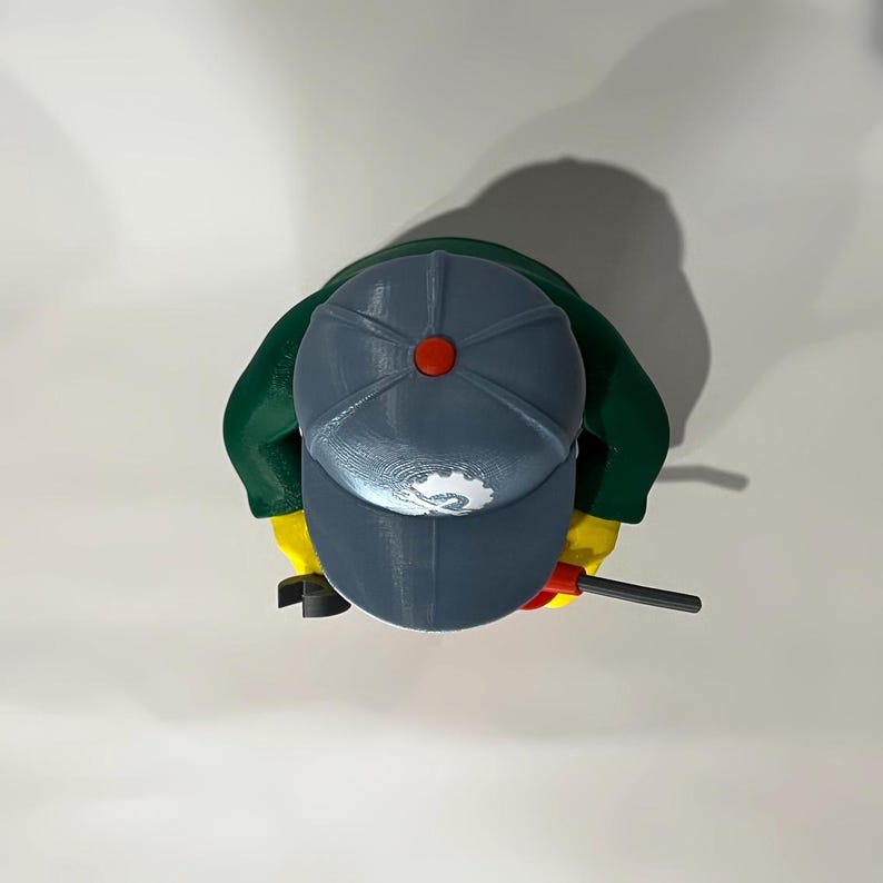 May include: Overhead view of a Lego figure wearing a gray cap with a red button, green jacket, and yellow hands. The figure holds a black and red tool. The cap has a white logo.